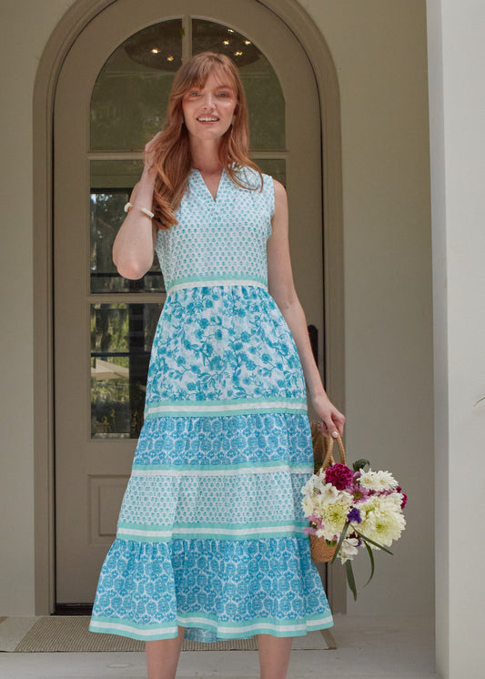 A redhead woman wearing the Cabana Life sun protective Rosebay Sleeveless Tiered Maxi Dress and holding a bag of flowers in front of a house.
