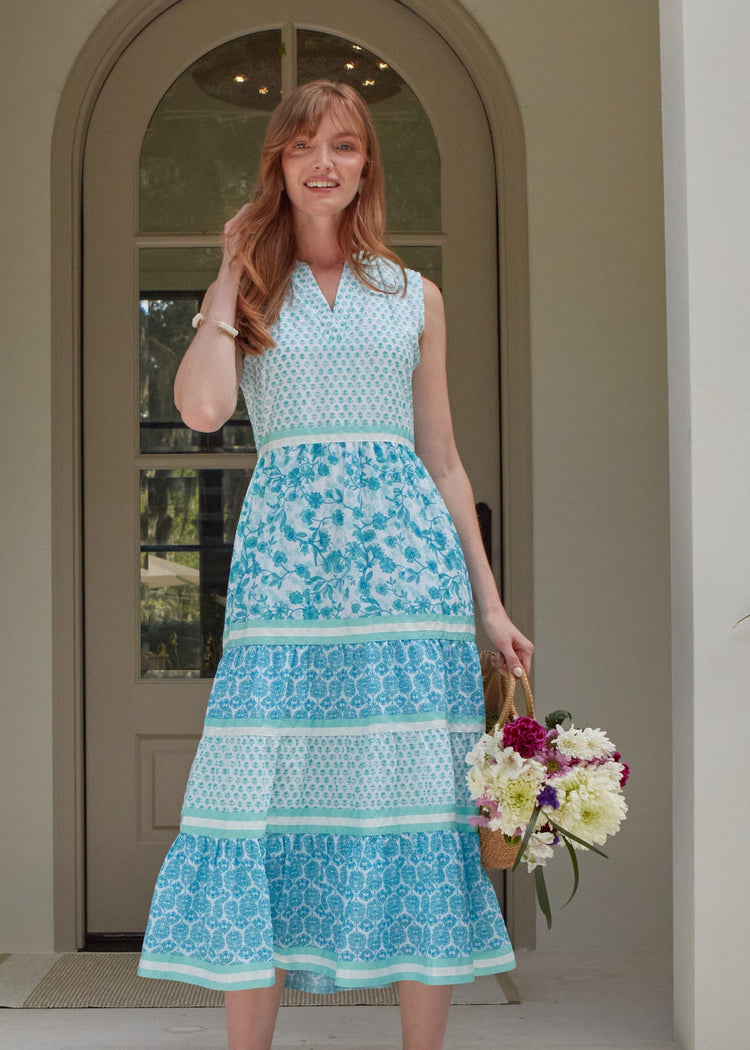 A redhead woman wearing the Cabana Life sun protective Rosebay Sleeveless Tiered Maxi Dress and holding a bag of flowers in front of a house.