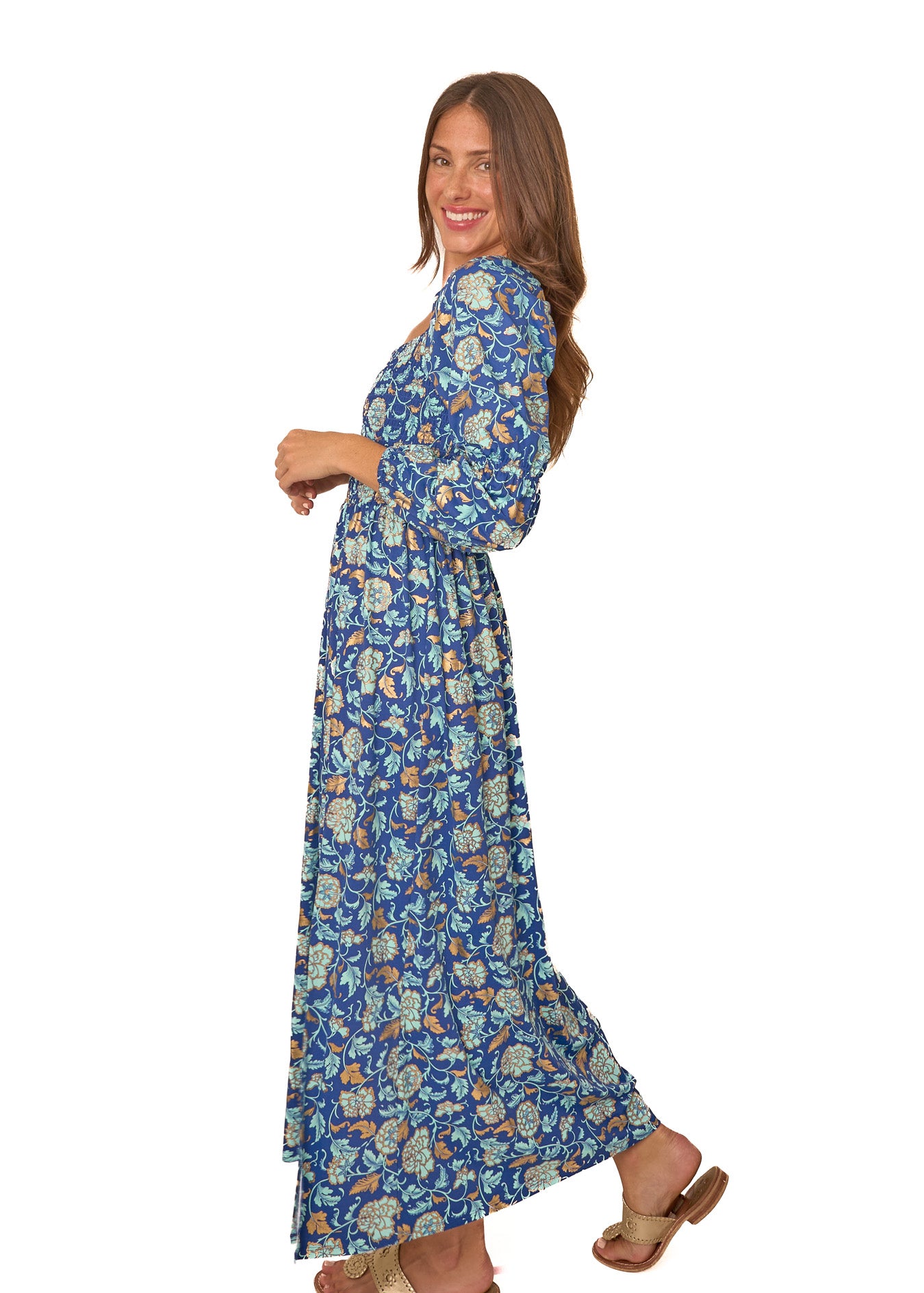 The side of a brunette woman wearing Cabana Life's sun protective Navy Floral Metallic Puff Sleeve Maxi Dress with gold shoes on a white background.