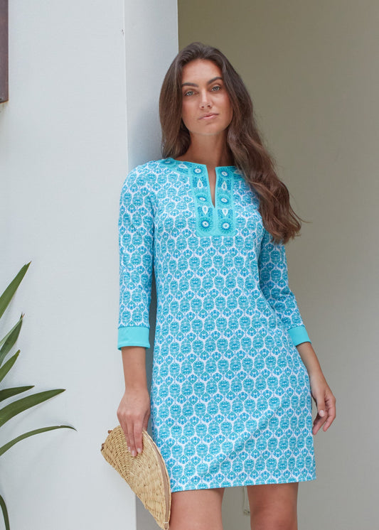 A brunette woman holding a rattan clutch and leaning against a white wall wearing the Cabana Life sun protective Rosebay Embroidered Tunic Dress.