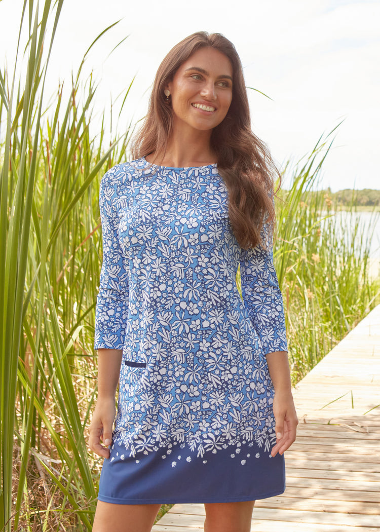 Woman wearing a blue floral dress standing in a natural setting with tall grass and water.