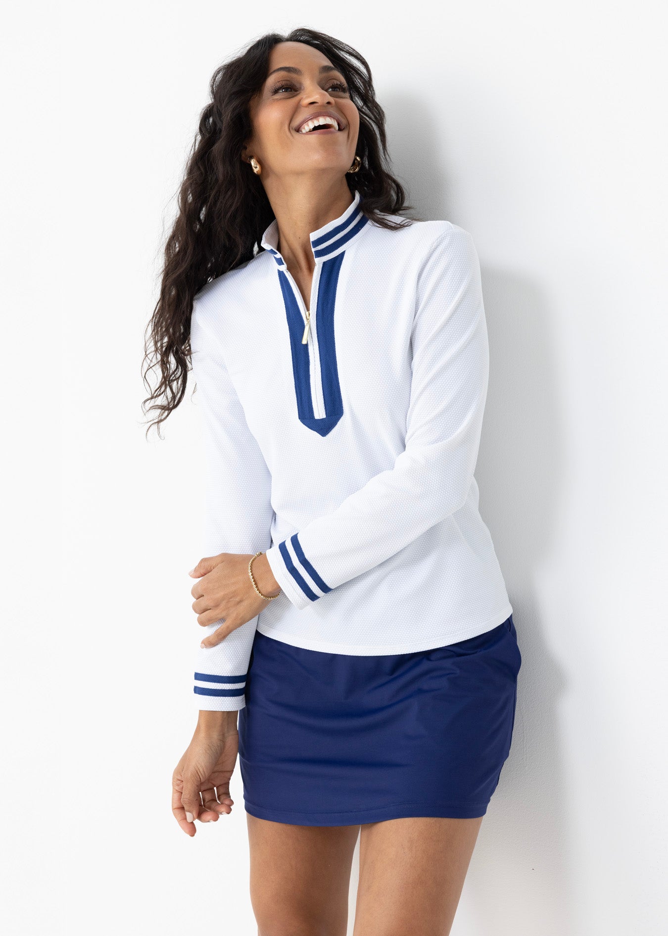 A brunette woman wearing the Cabana Life White Collared 1/4 Zip with the Navy 15" Skort in a studio laughing.