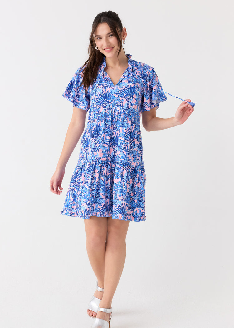 A brunette woman holding the tassel tie on the Cabana Life sun protective Palm Beach Short Sleeve Tiered Dress, paired with silver sandals.