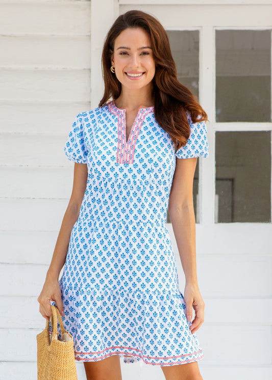 Woman wearing a blue patterned dress standing in front of a white wall.