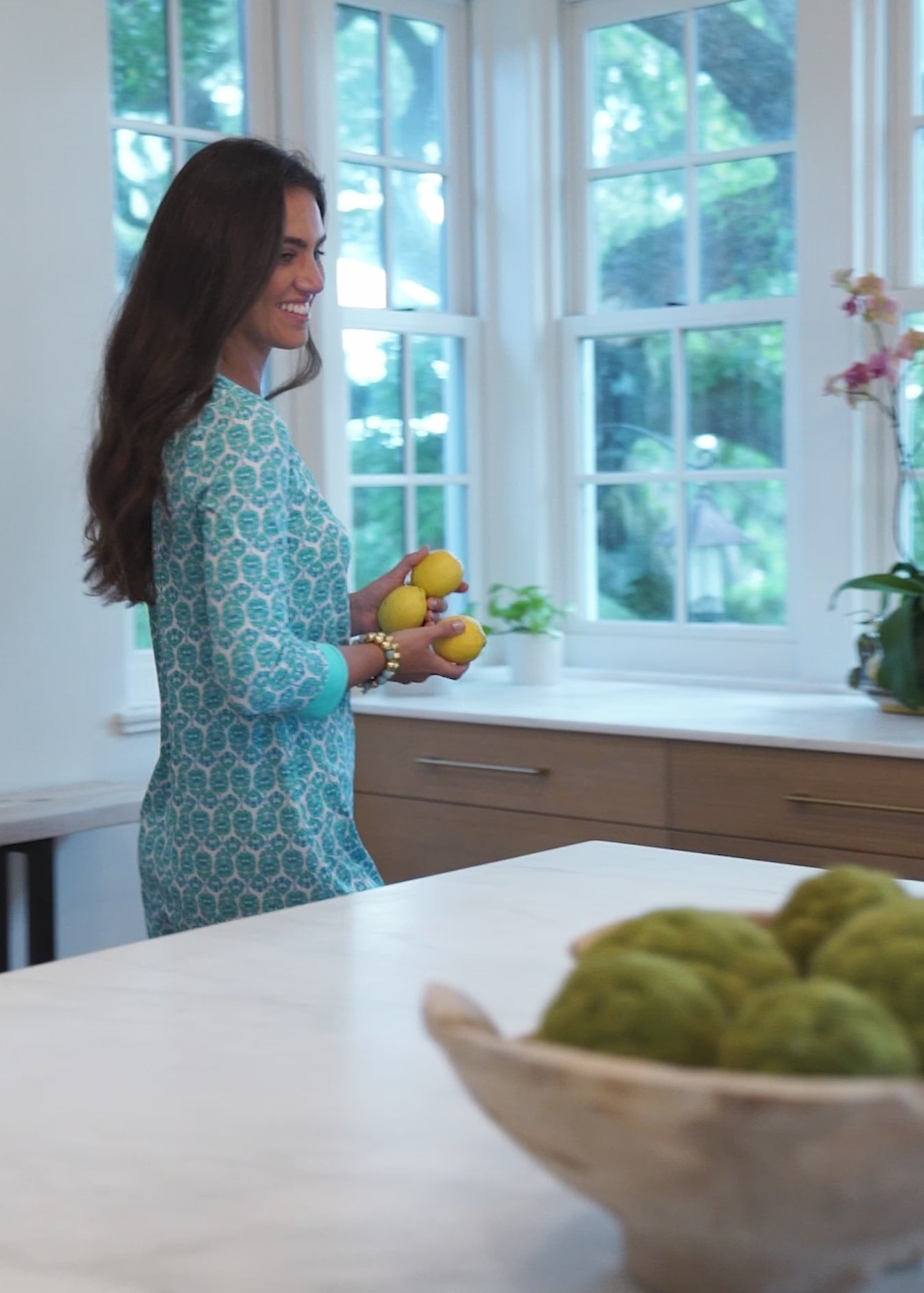 A brunette woman making lemonade inside while wearing the Cabana Life sun protective Rosebay Embroidered Tunic Dress.