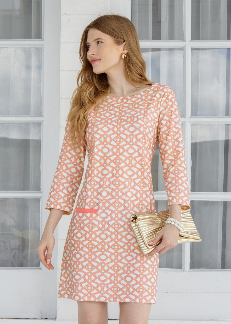 Woman wearing a patterned dress holding a gold clutch in front of a window.