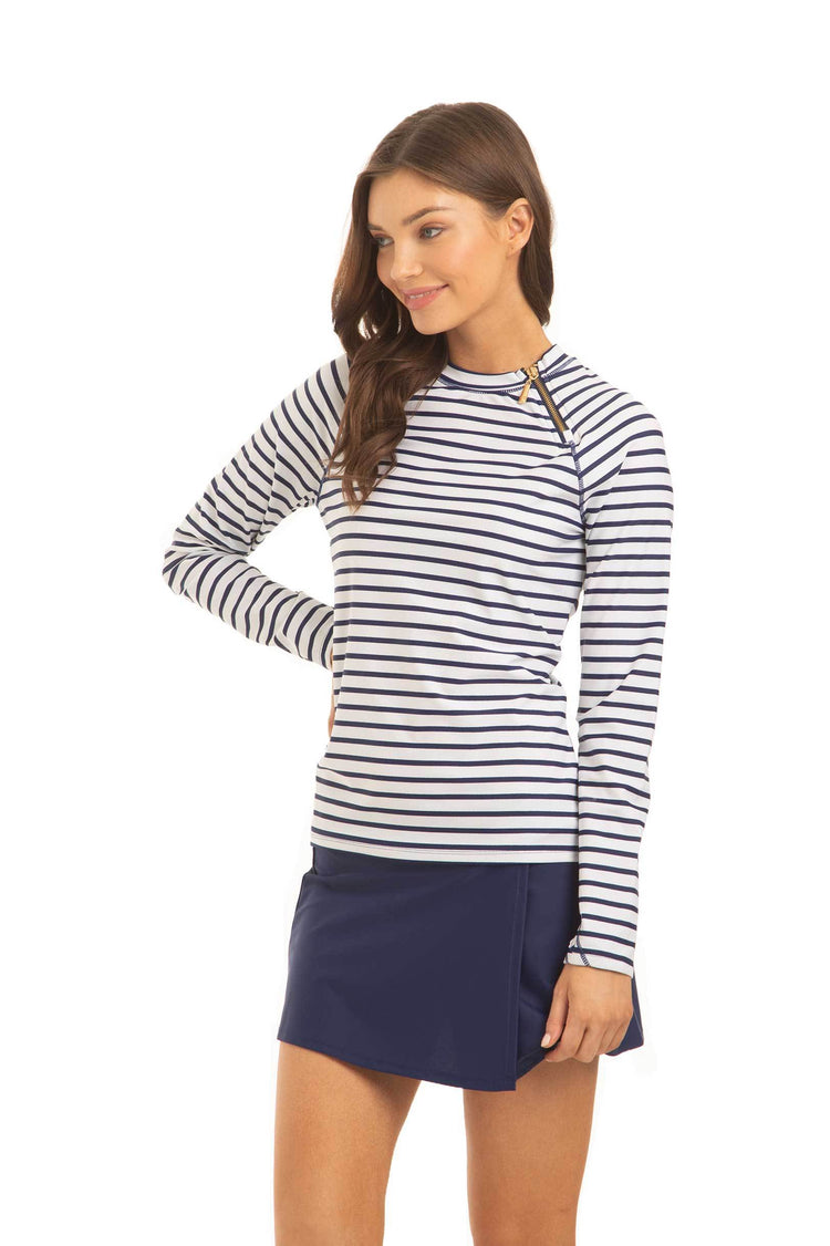 A brunette woman with her hand on her  hip facing the side and wearing the Cabana Life sun protective Navy Stripe Zipper Rashguard and Navy Classic Swim Skirt.