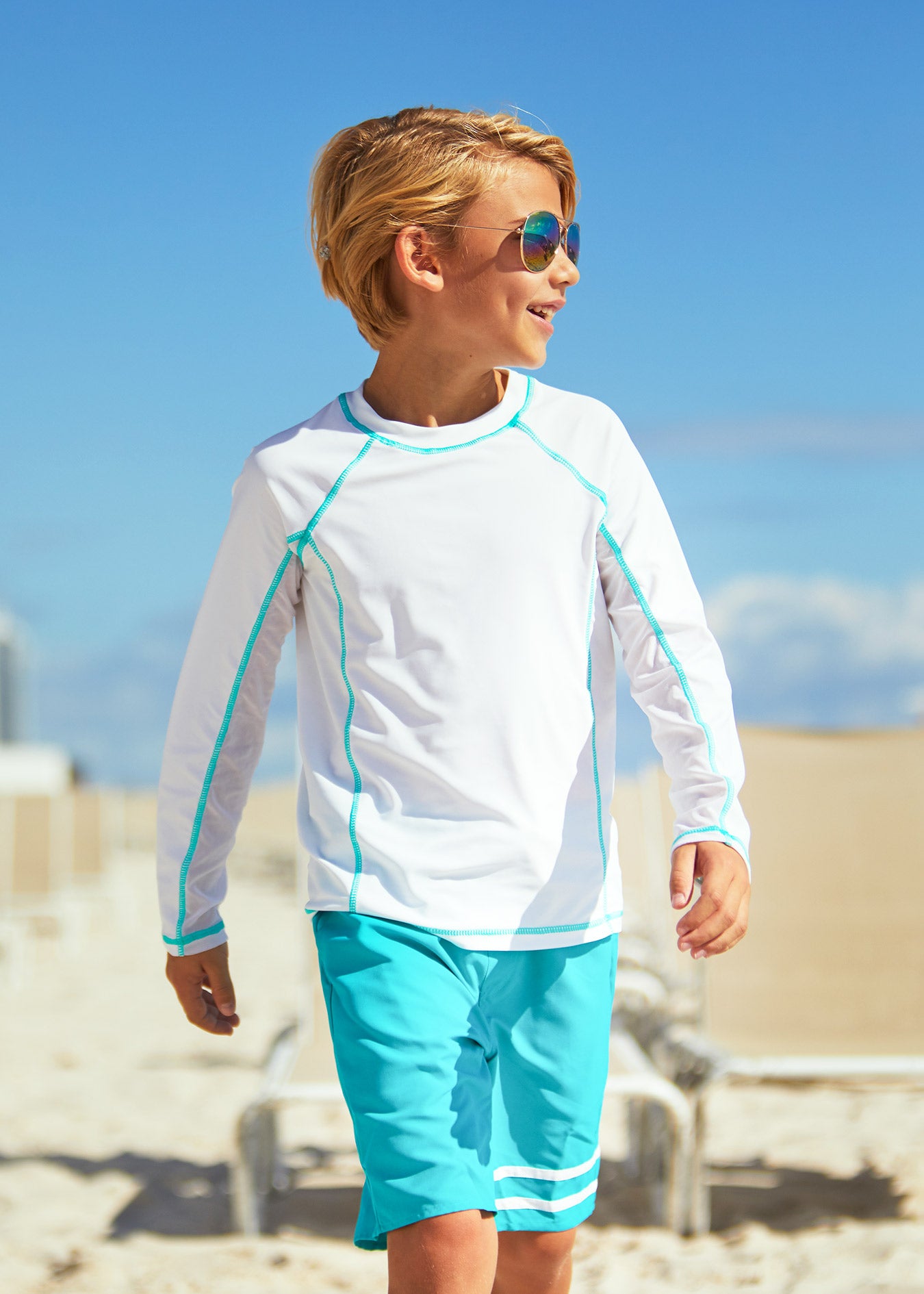 Boy walking on beach wearing Cabana Life Boys White Long Sleeve Rashguard and Boys Turquoise Swim Trunks 