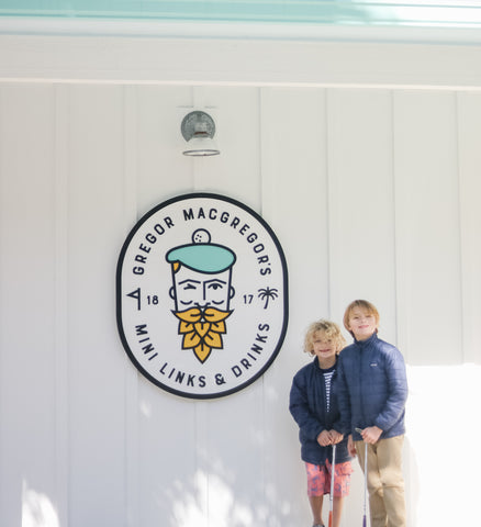 Two boys infront of Gregor Mcgregor's Mini Links & Drinks sign wearing Boy's Red Crab Swim Shorts and Rashguard set