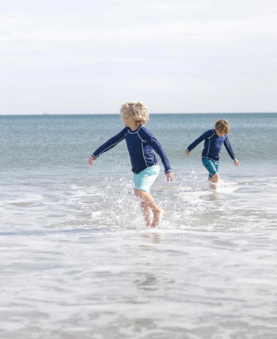 Two boys running on beach wearing boys Navy Long Sleeve Rashguards with Aruba Blues Swim Trunks and Coral Tides Swim Trunks