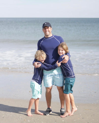 Man wearing Men's Navy Long Sleeve Rashguard with Coastal Cottage Swim Trunks and boy wearing Boy's Navy Rashguard with Coral Tides Swim Trunks and boy wearing Boy's Navy Rashguard with Boy's Aruba Blues Swim Trunks